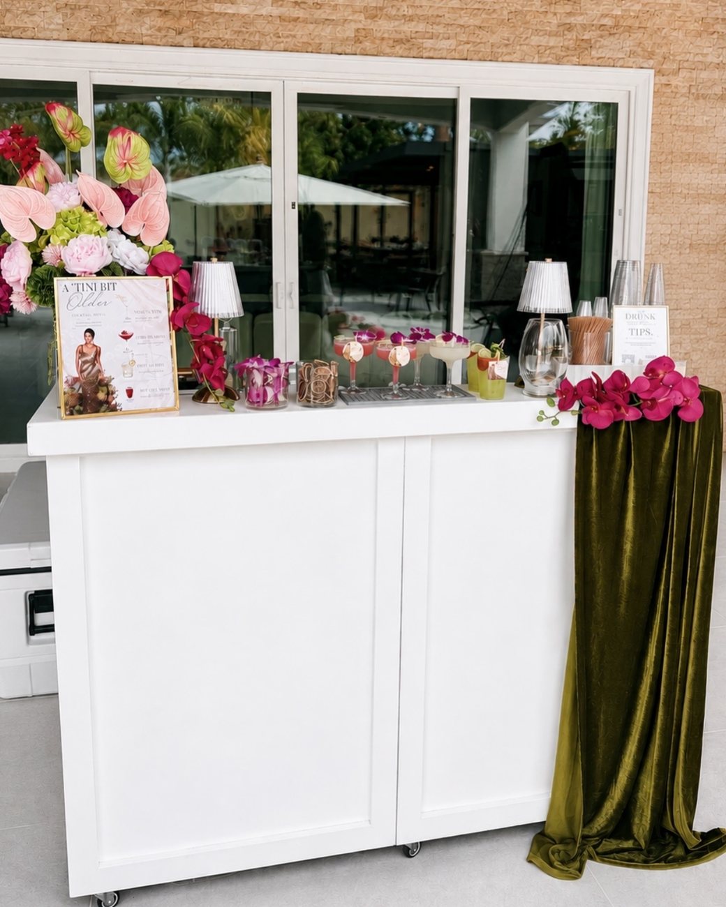 White mobile bar with pink orchids, glowing lamps, a custom cocktail menu and a draped olive velvet runner