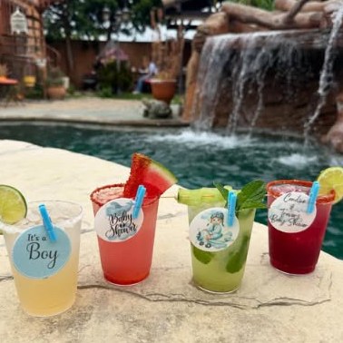 Four cocktails with custom toppers lined up beside a pool and waterfall