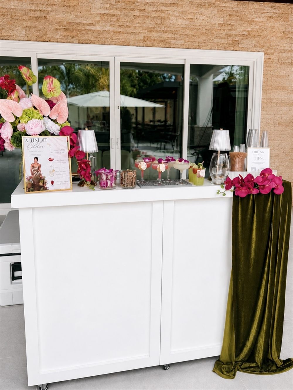 White mobile bar styled with pink florals, orchids, a velvet green runner and a custom cocktail menu